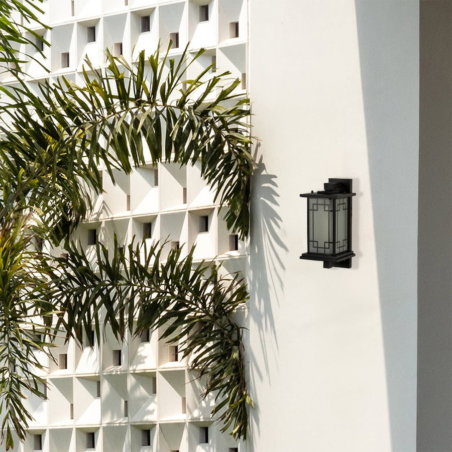 Gold outdoor wall lantern with the light off installed on a bright exterior wall surrounded by green plants enhancing elegant outdoor architectural styling.