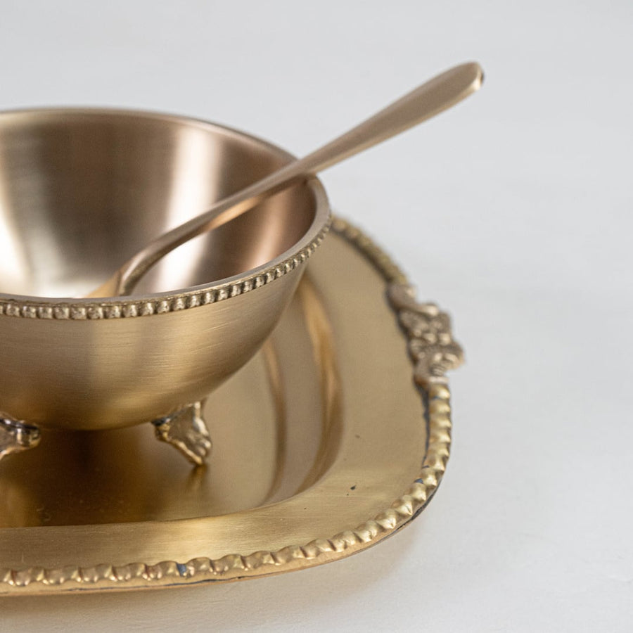 Closeup image of gold metal condiment bowl with decorative tray and spoon for elegant table presentation decor.