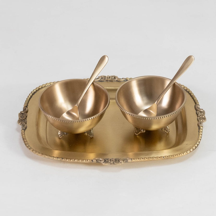 Twin gold serving bowls with spoons placed symmetrically on decorative rectangular gold tray on white background.