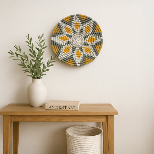 Handcrafted woven wall plate with green mustard and cream patterns displayed on a white background above a wooden console styled with books a ceramic vase and minimal decor for a calm lifestyle scene.