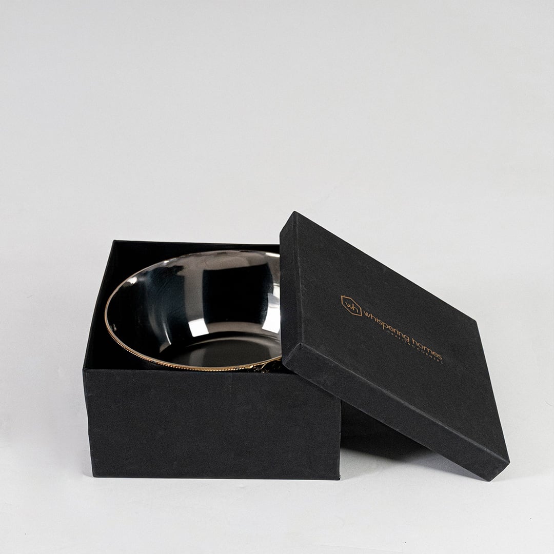 Round silver bowl with gold details shown partially packed in black branded gift box with lid slightly opened.