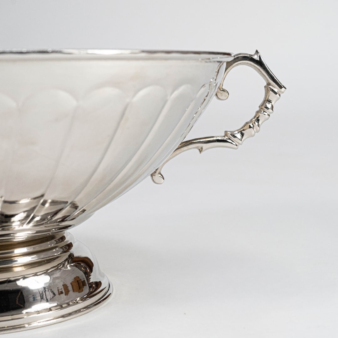 Side closeup of silver fruit bowl highlighting decorative curved handle and glossy scalloped exterior on white.