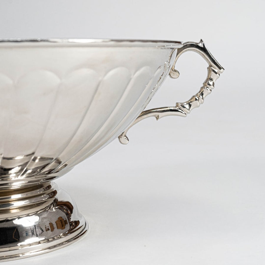 Side closeup of silver fruit bowl highlighting decorative curved handle and glossy scalloped exterior on white.
