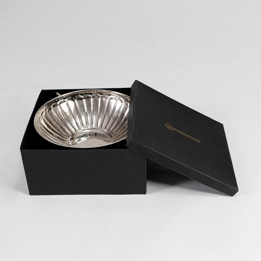 Silver metal fruit bowl partially placed inside luxury black gift box with logo branding shown on white backdrop.