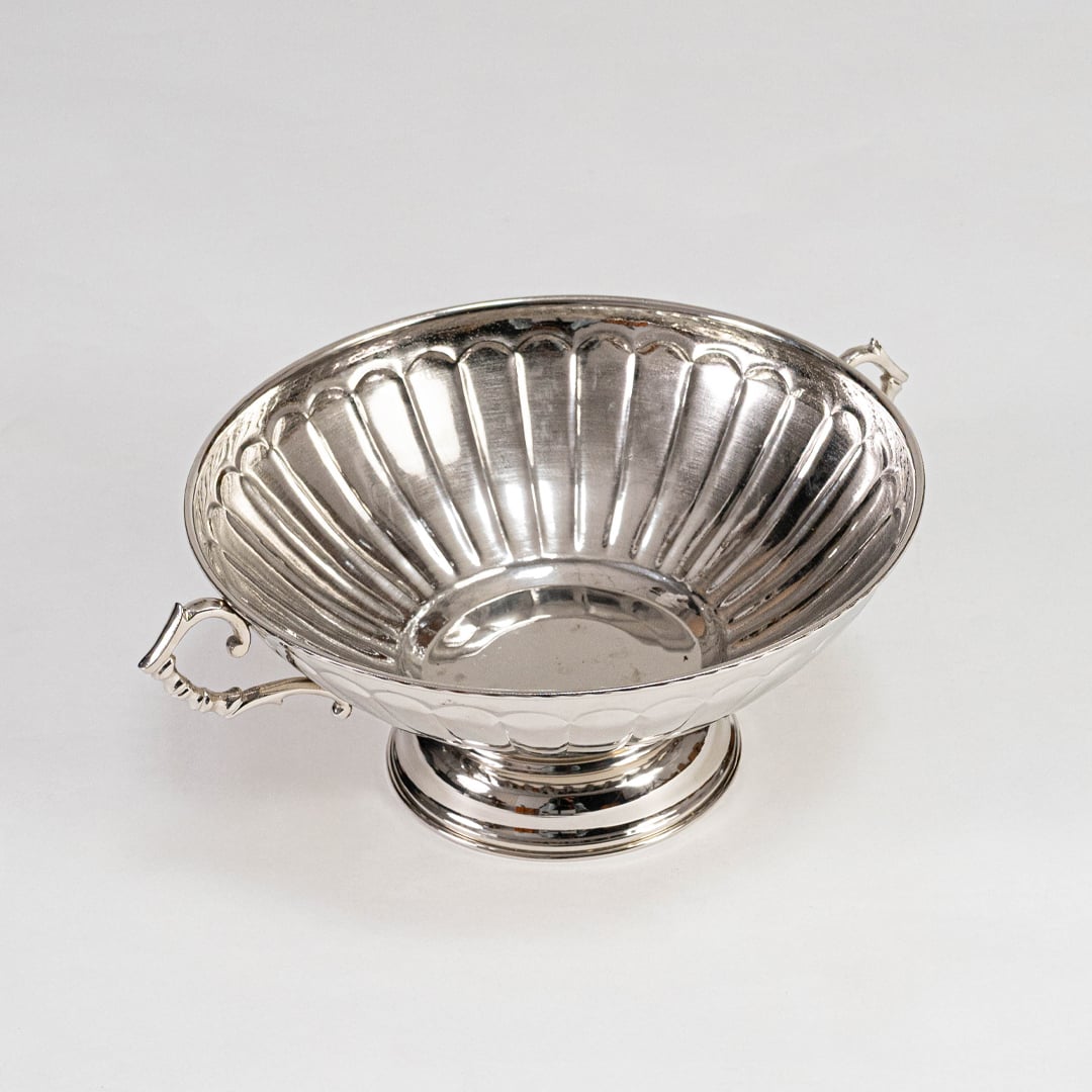 Shiny silver-toned fluted fruit bowl with curved handles on pedestal base placed on a plain white background.