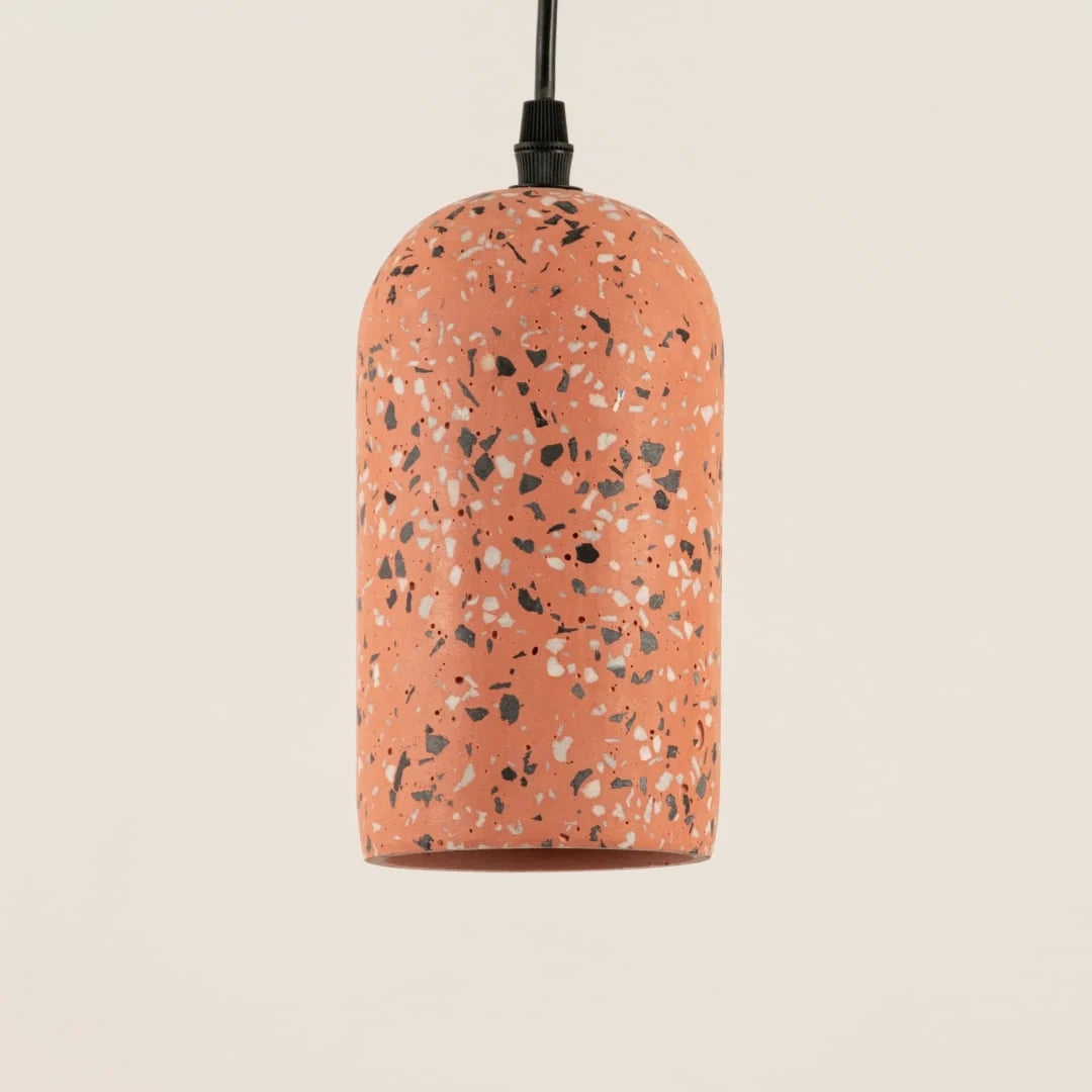 A terrazzo-patterned pendant light with a rounded cylindrical shade in terracotta orange, speckled with black, white, and gray chips, suspended by a black cord against a light beige background.