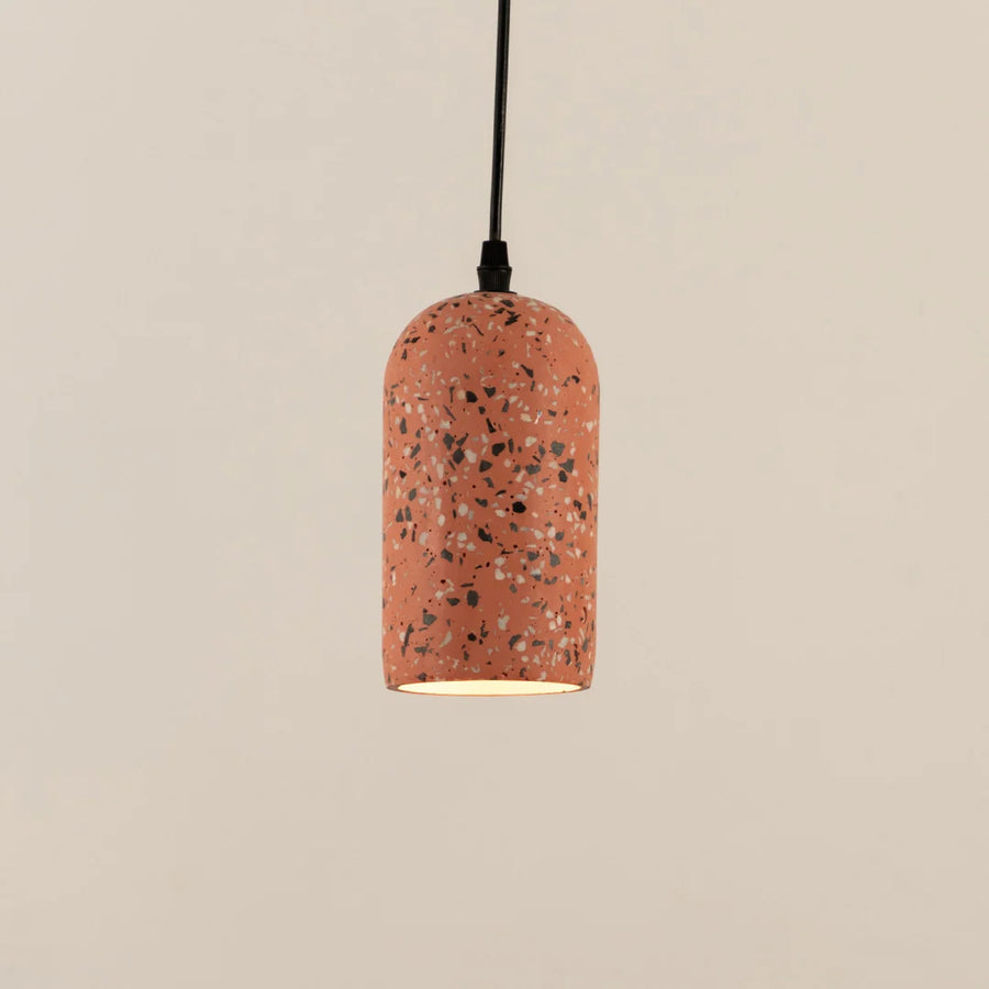 A terrazzo-patterned pendant light with a cylindrical shade suspended by a black cord against a warm beige background.