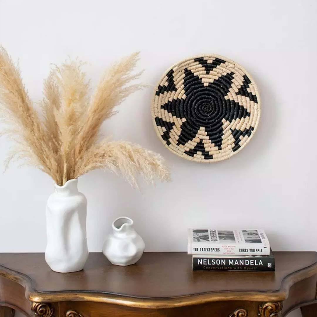 Decorative setup with a woven wall basket, pampas grass in a vase, and books on a wooden surface.