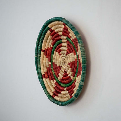 Round jute woven wall basket decor in beige red and green rustic boho handmade spiral wall art accent piece side angle view.