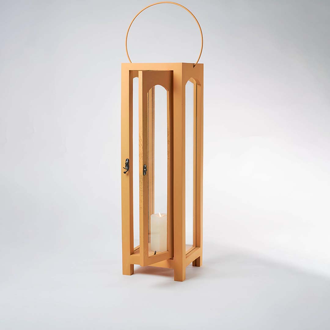 Illuminated wooden lantern with a lit white pillar candle inside against a plain white background showing the warm glow through glass panes.