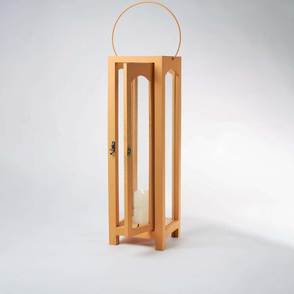 Illuminated wooden lantern with a lit white pillar candle inside against a plain white background showing the warm glow through glass panes.