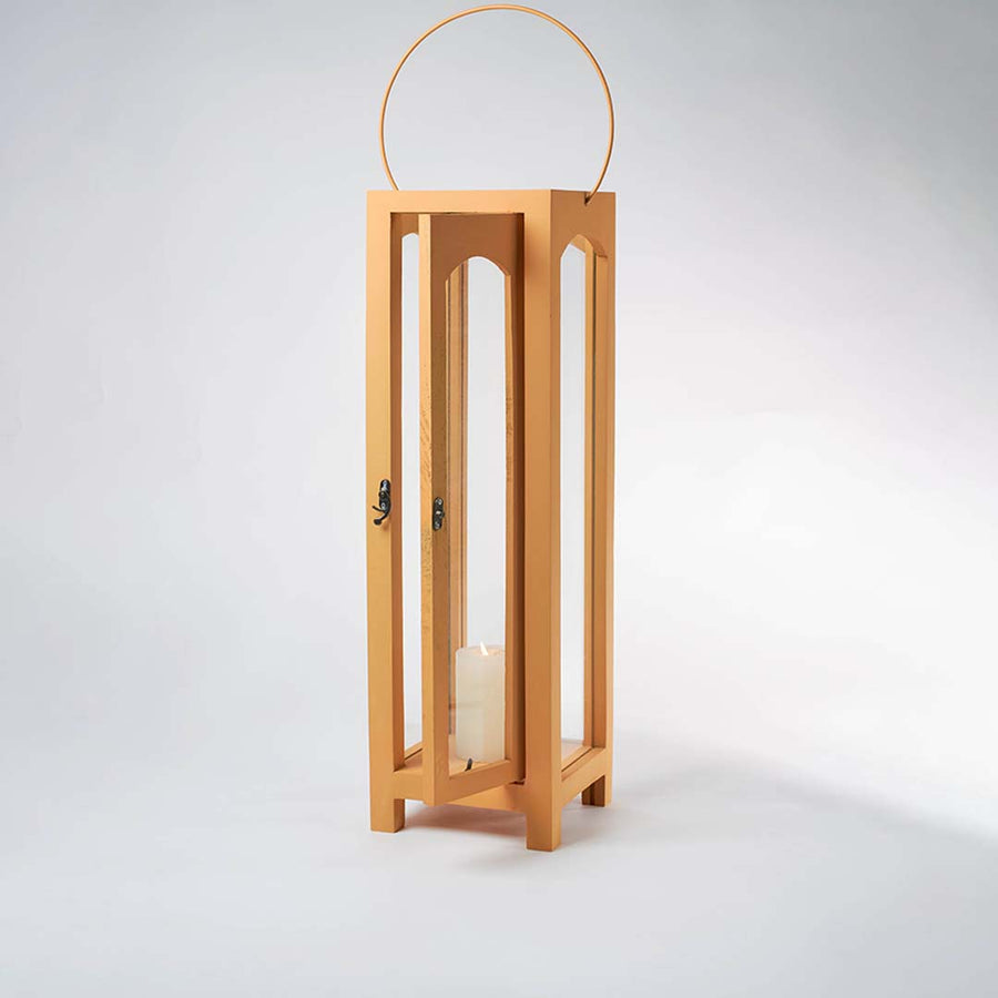 Illuminated wooden lantern with a lit white pillar candle inside against a plain white background showing the warm glow through glass panes.
