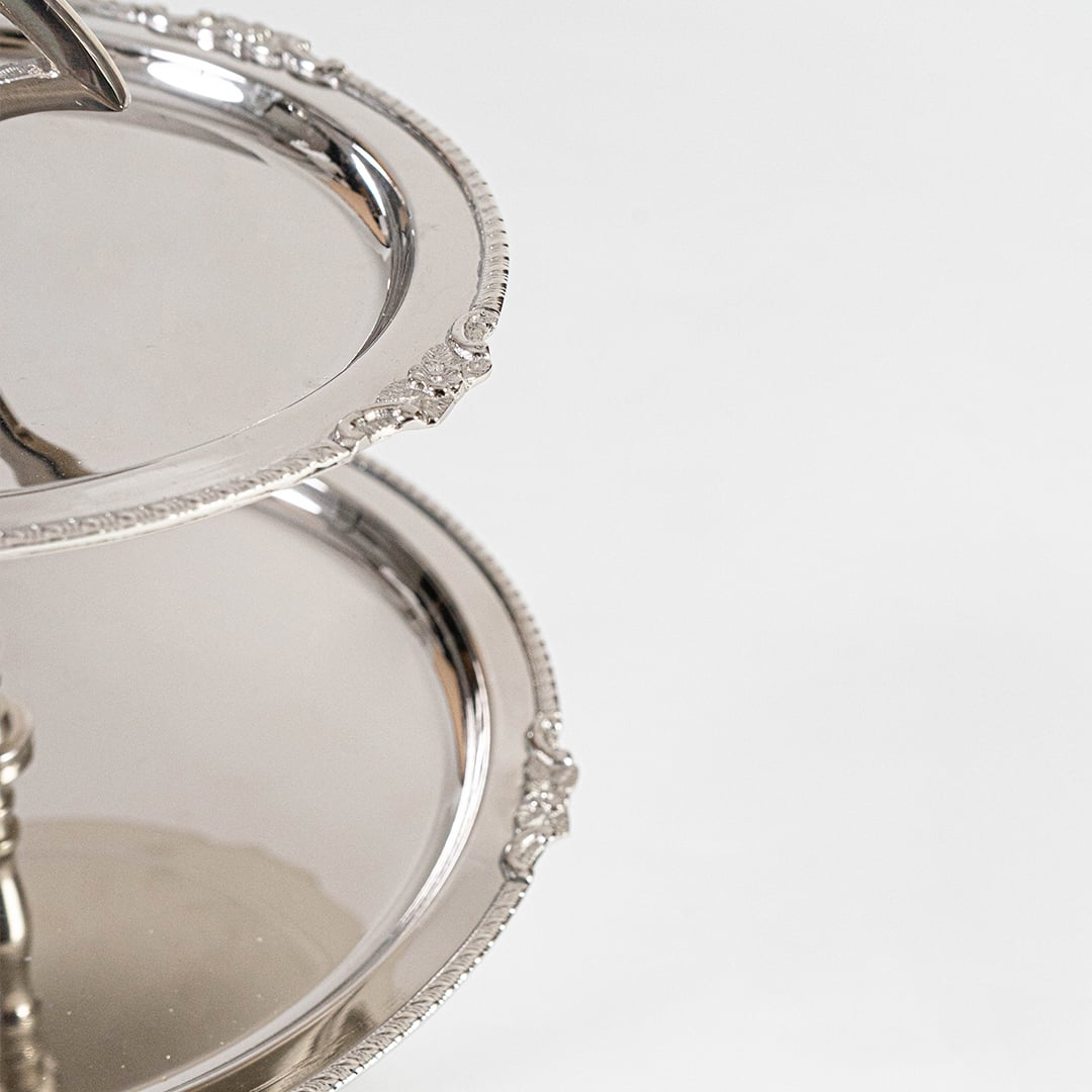 Silver two tier metal serving tray with sweets and fruit displayed on a beige tablecloth for elegant home entertaining.