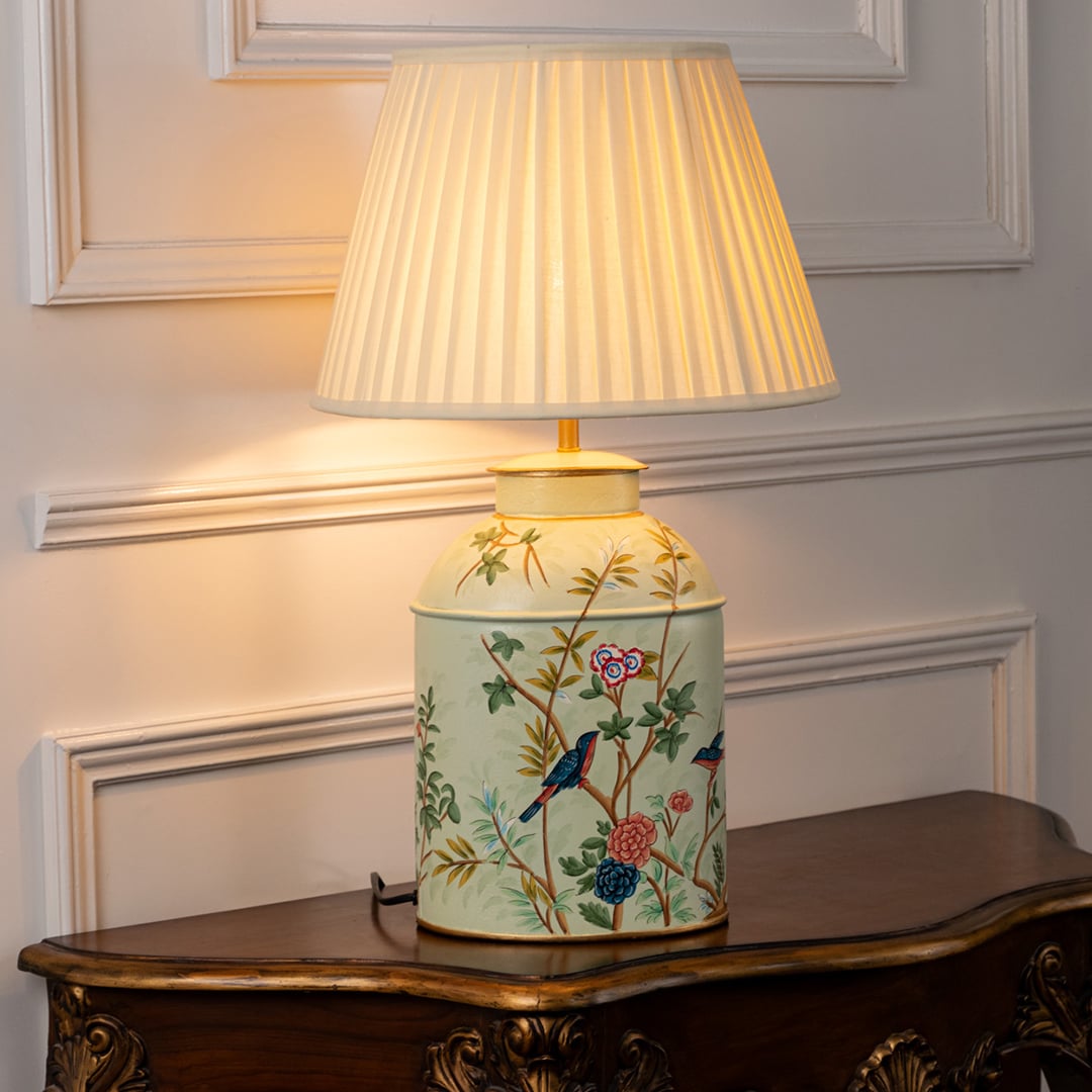 Table lamp with white pleated fabric shade and cream ceramic base featuring hand painted floral artwork styled on a wooden table against a white background.