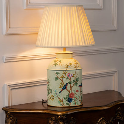 Table lamp with white pleated fabric shade and cream ceramic base featuring hand painted floral artwork styled on a wooden table against a white background.