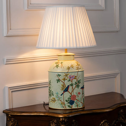 Table lamp with white pleated fabric shade and cream ceramic base featuring hand painted floral and bird design placed on a wooden table against a white background.