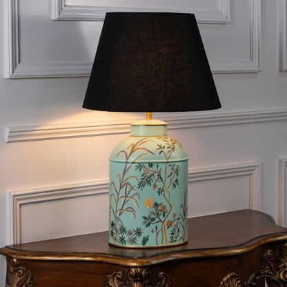 Table lamp with black fabric shade and light turquoise ceramic base decorated with floral patterns styled on a wooden table against a white background.