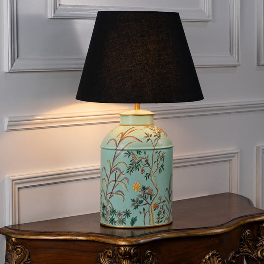 Table lamp with black fabric shade and light turquoise ceramic base decorated with floral patterns styled on a wooden table against a white background.