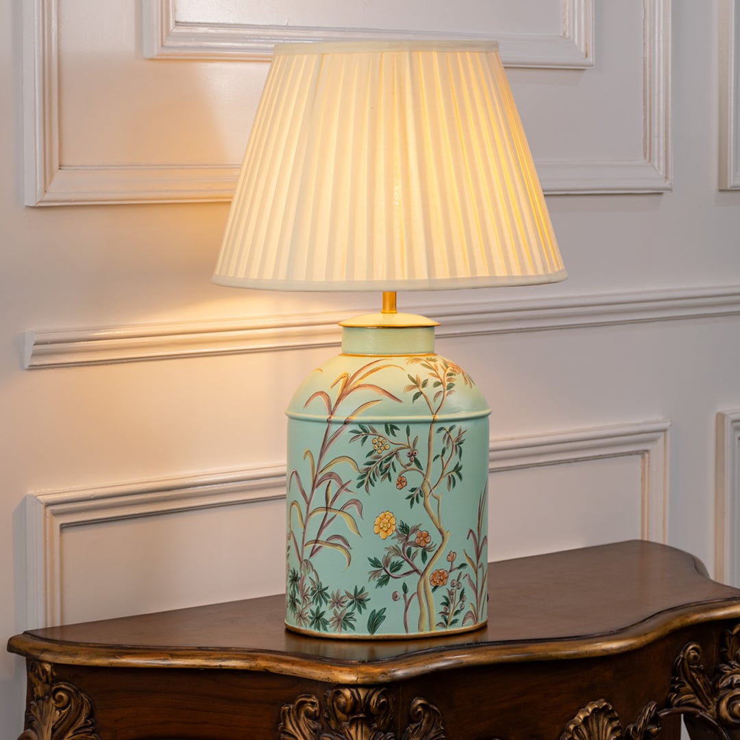 Table lamp with white pleated fabric shade and light blue ceramic base decorated with delicate floral patterns placed on a wooden table against a white background.