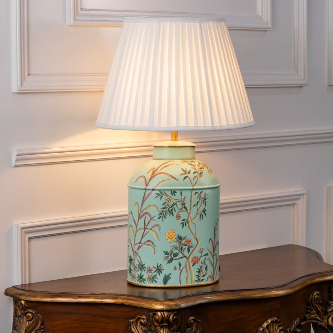 Table lamp with white pleated fabric shade and pastel blue ceramic base decorated with elegant floral patterns displayed on a wooden table against a white background.