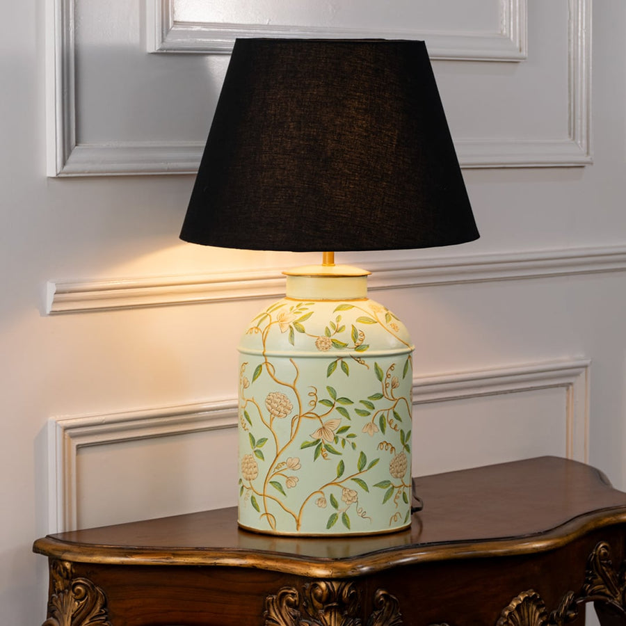 Table lamp with black fabric shade and off white ceramic base with delicate green leaf patterns placed on a wooden table against a white background.