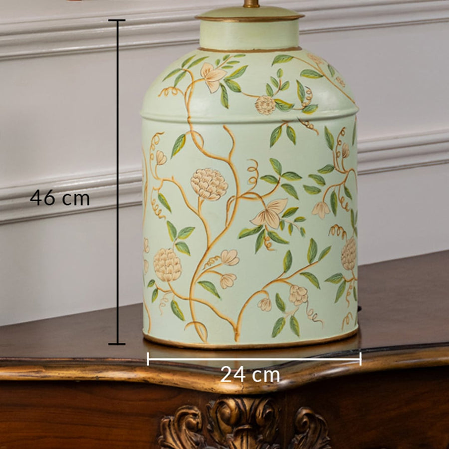 Table lamp with white pleated fabric shade and off white ceramic base featuring soft green botanical detailing shown on a wooden table against a white background.