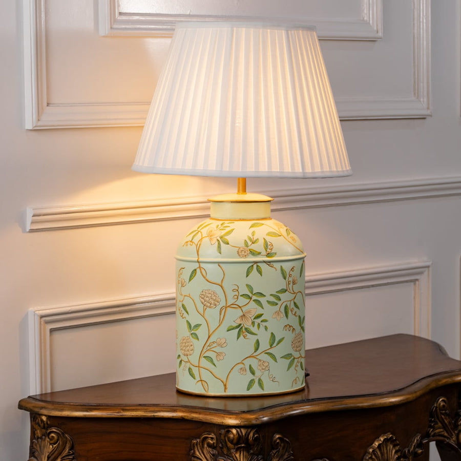 Table lamp with white pleated fabric shade and off white ceramic base featuring soft green botanical motifs styled on a wooden table against a white background.