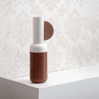 Tall white and brown ribbed ceramic vase accented with a decorative wood textured half disc against damask wallpaper.
