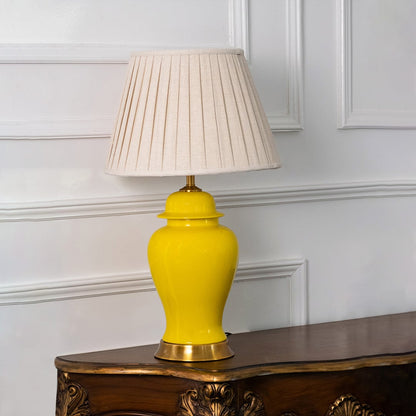 Yellow ceramic lamp with golden base and fabric pleated shade displayed against white background.