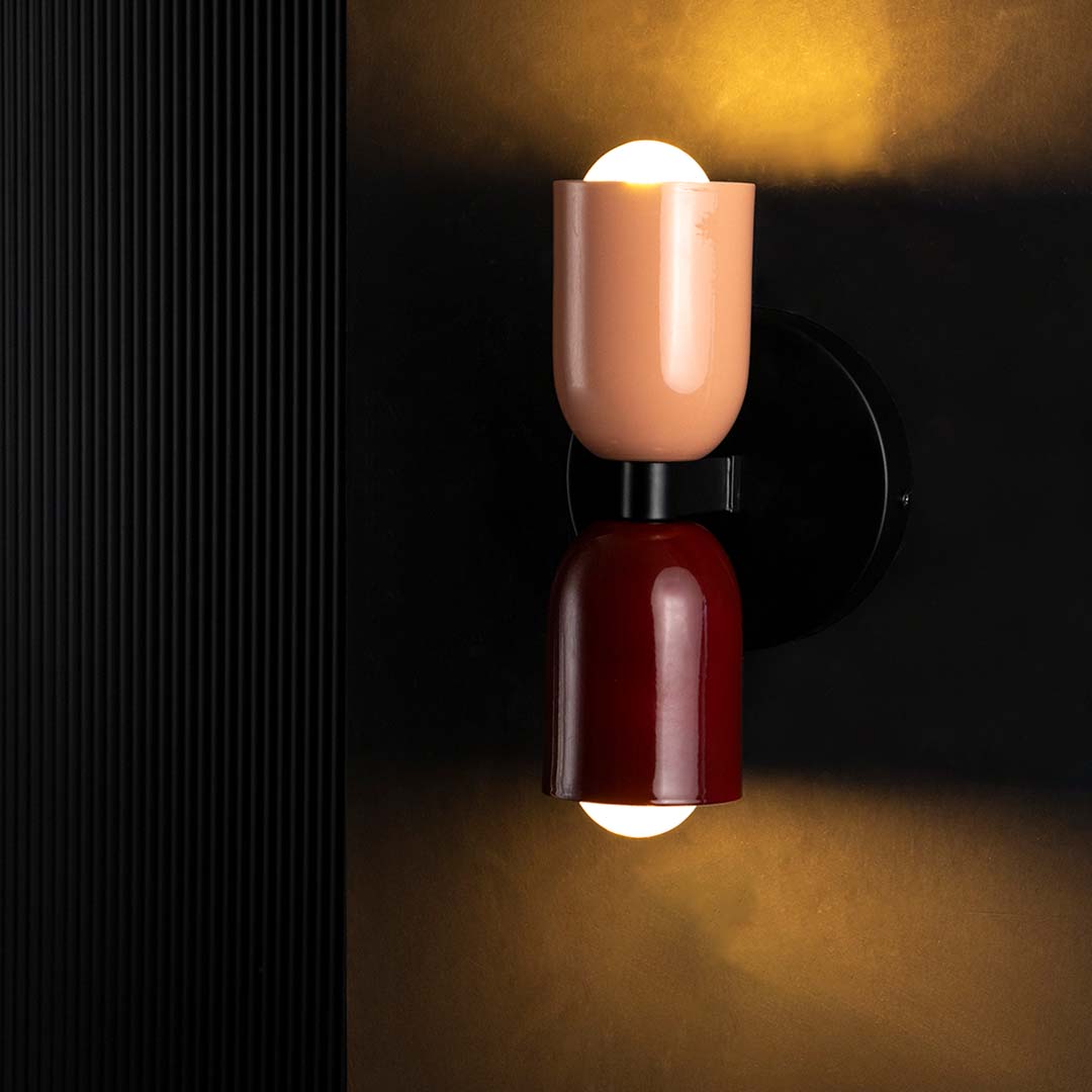 A contemporary dual tone wall light glowing warmly on a black background with the top bulb lit creating a soft halo around the maroon and peach shades.