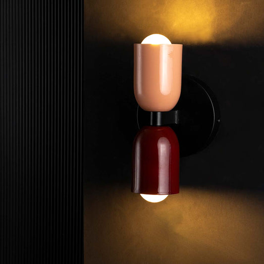 A contemporary dual tone wall light glowing warmly on a black background with the top bulb lit creating a soft halo around the maroon and peach shades.