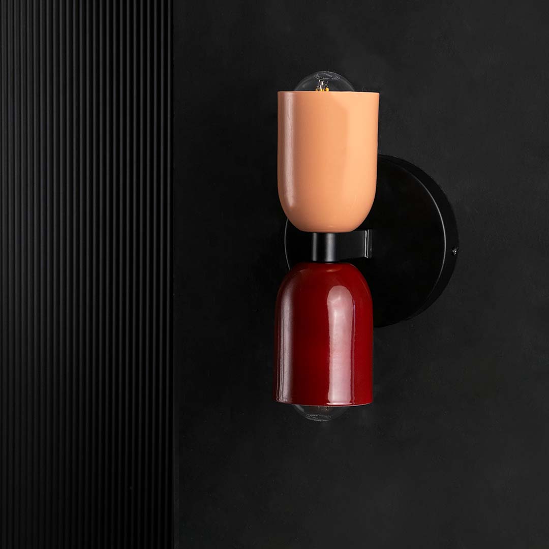 A modern wall sconce with maroon and peach shades shown against a black background with both bulbs turned off emphasizing its glossy finish and sleek form.