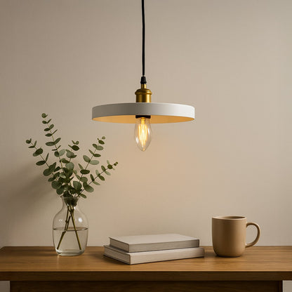 Modern white ring pendant light glowing warmly above a wooden table with books a mug and a glass vase with green leaves.