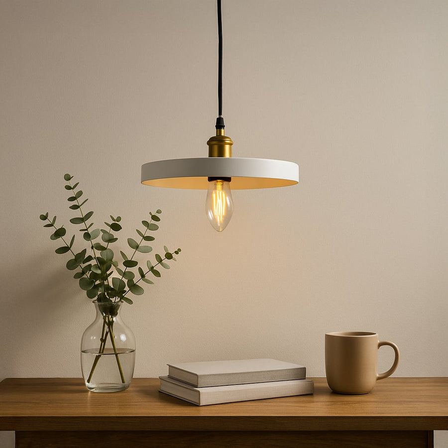 Modern white ring pendant light glowing warmly above a wooden table with books a mug and a glass vase with green leaves.