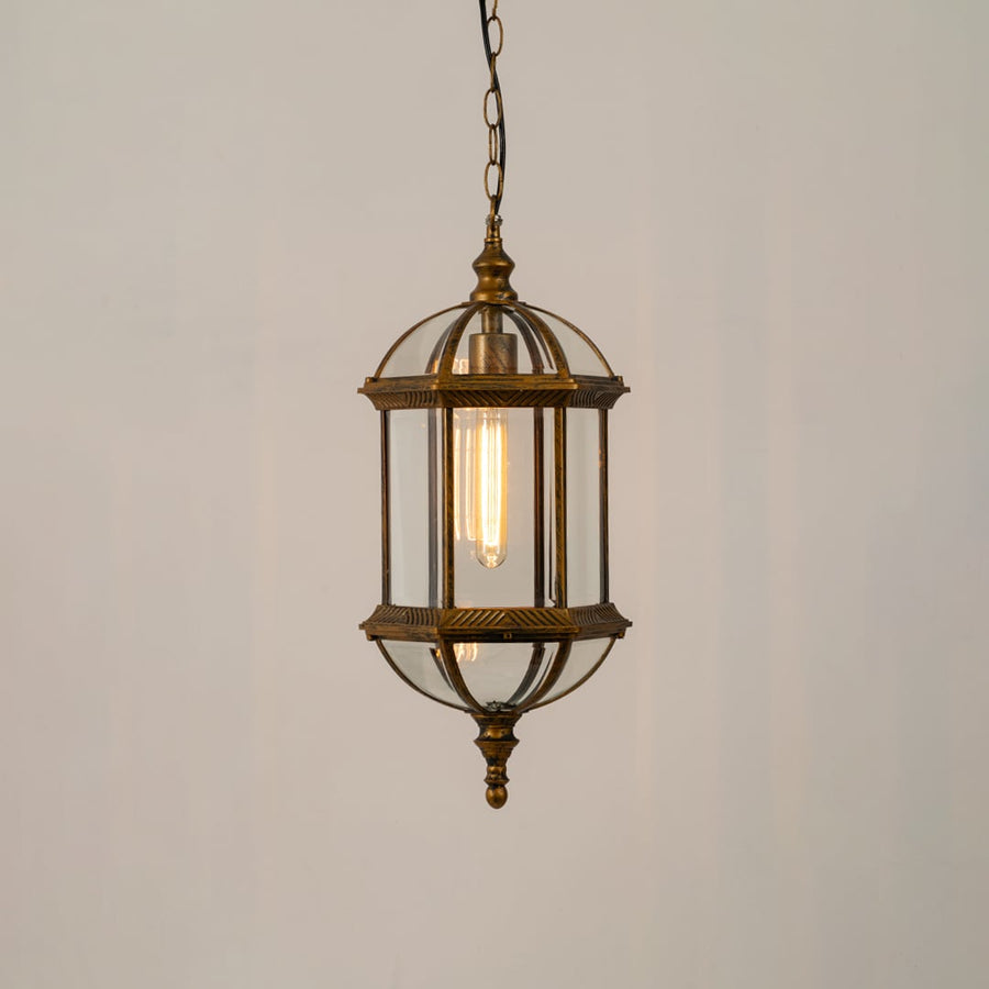 A bronze vintage style hanging lantern lit with a warm glow showcasing its elegant glass panels and dome top design suspended from a chain against a neutral wall backdrop.