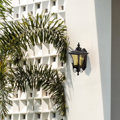 Bronze metal outdoor wall light lifestyle image with amber glass shade installed on a white villa facade for decor.