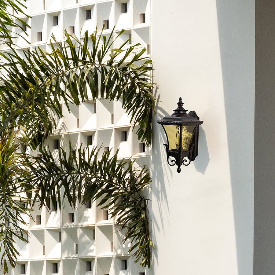 Bronze metal outdoor wall light lifestyle image with amber glass shade installed on a white villa facade for decor.