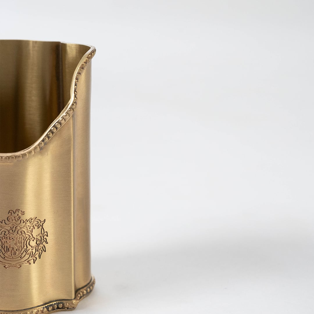 Closeup detail of tall gold metal pen holder showing engraved crest and scalloped edge against white background.