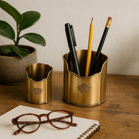 Elegant gold pencil holders displayed on a wooden desk with pens pencils and glasses placed beside a plant creating a warm organized workspace atmosphere.