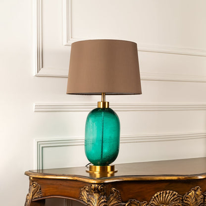 Green glass lamp with brown shade on carved console table with light off and white panelled wall for modern elegant décor.