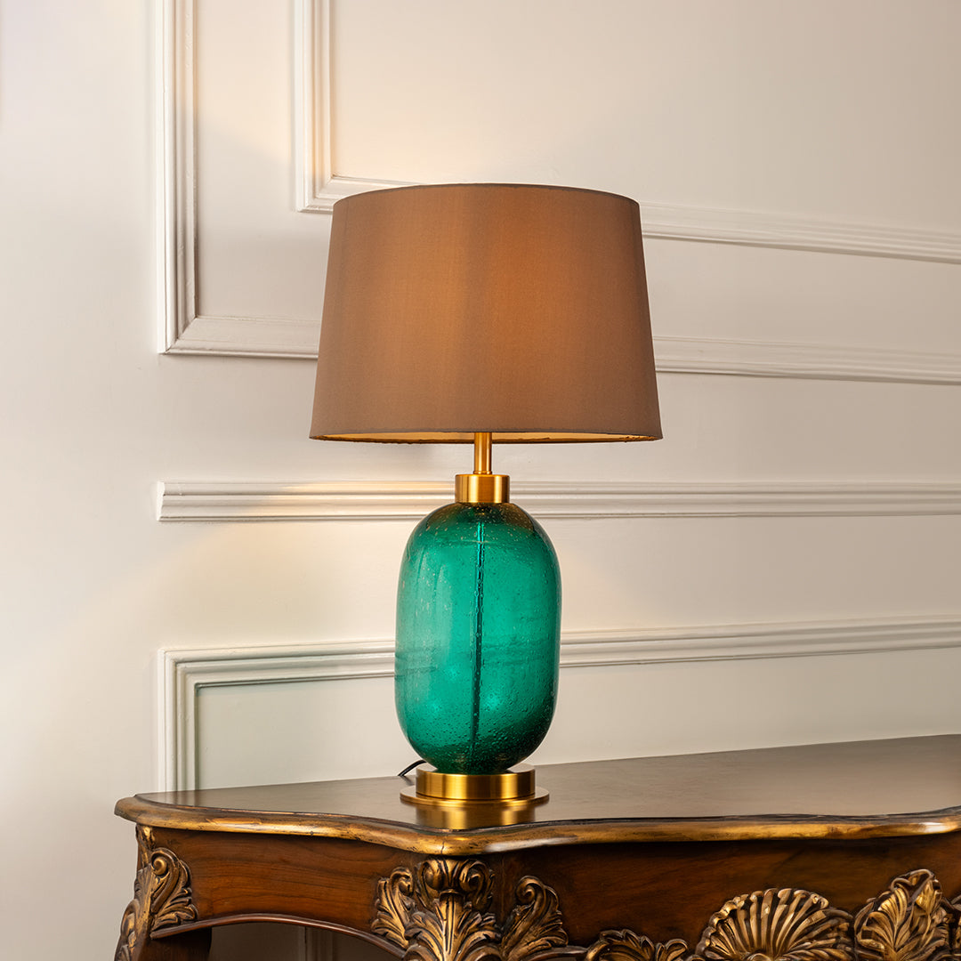 Turquoise glass table lamp with brown fabric shade displayed on a carved wooden console table creating warm lighting.