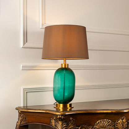 Turquoise glass table lamp with brown fabric shade displayed on a carved wooden console table creating warm lighting.