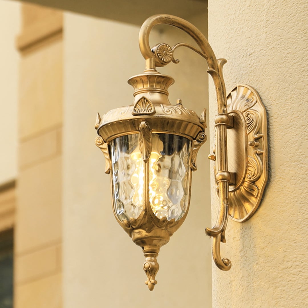 A gold antique outdoor wall lantern with the light on illuminating the textured wall and creating a warm elegant glow that enhances the classic architectural exterior.