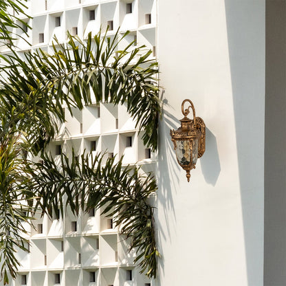 A gold traditional outdoor wall lantern with the light off mounted on a modern white exterior wall surrounded by palm leaves showcasing its ornate curved arm and detailed metalwork.