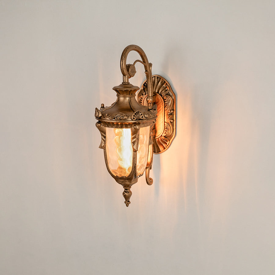Antique brass metal outdoor wall light on white background with ornate scroll accents and clear glass shade for decor.