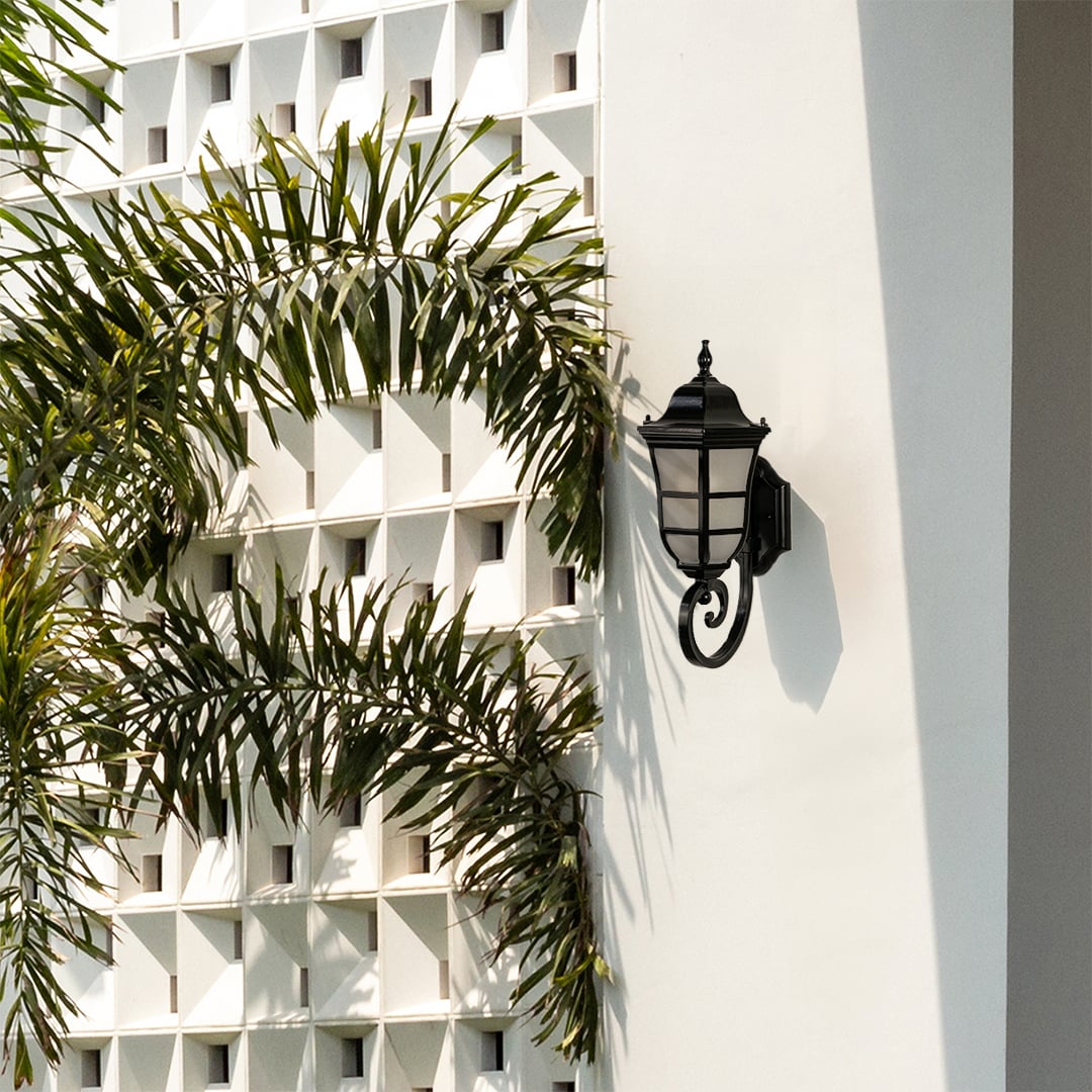 Classic black metal outdoor wall lantern light off with frosted glass mounted on a modern exterior wall surrounded by palm leaves ideal for patio porch and garden Decor.