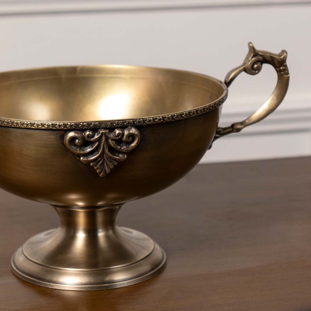 Closeup of antique brass pedestal bowl with ornate detailing and handles perfect for table centerpiece and luxury home decor.