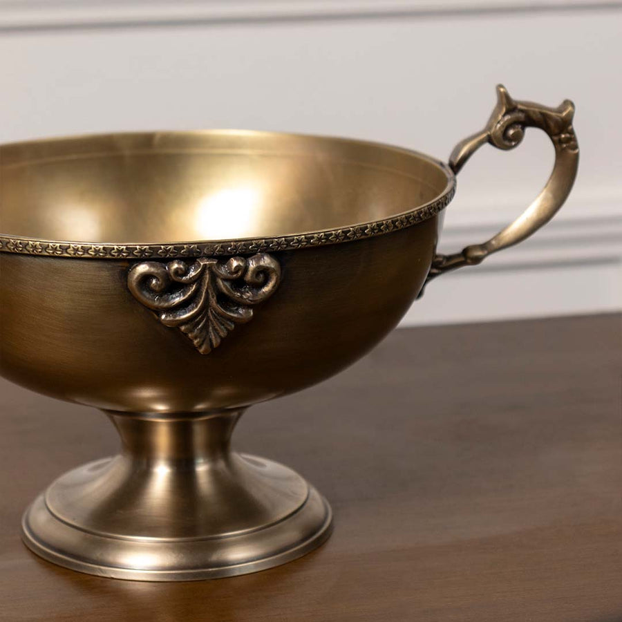 Closeup of antique brass pedestal bowl with ornate detailing and handles perfect for table centerpiece and luxury home decor.