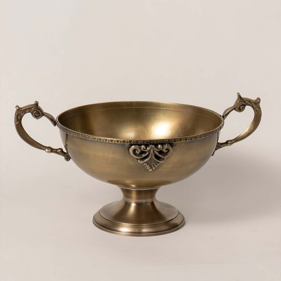 Vintage brass bowl with ornate handles placed on white surface against neutral background showcasing classic decorative home accent styling.