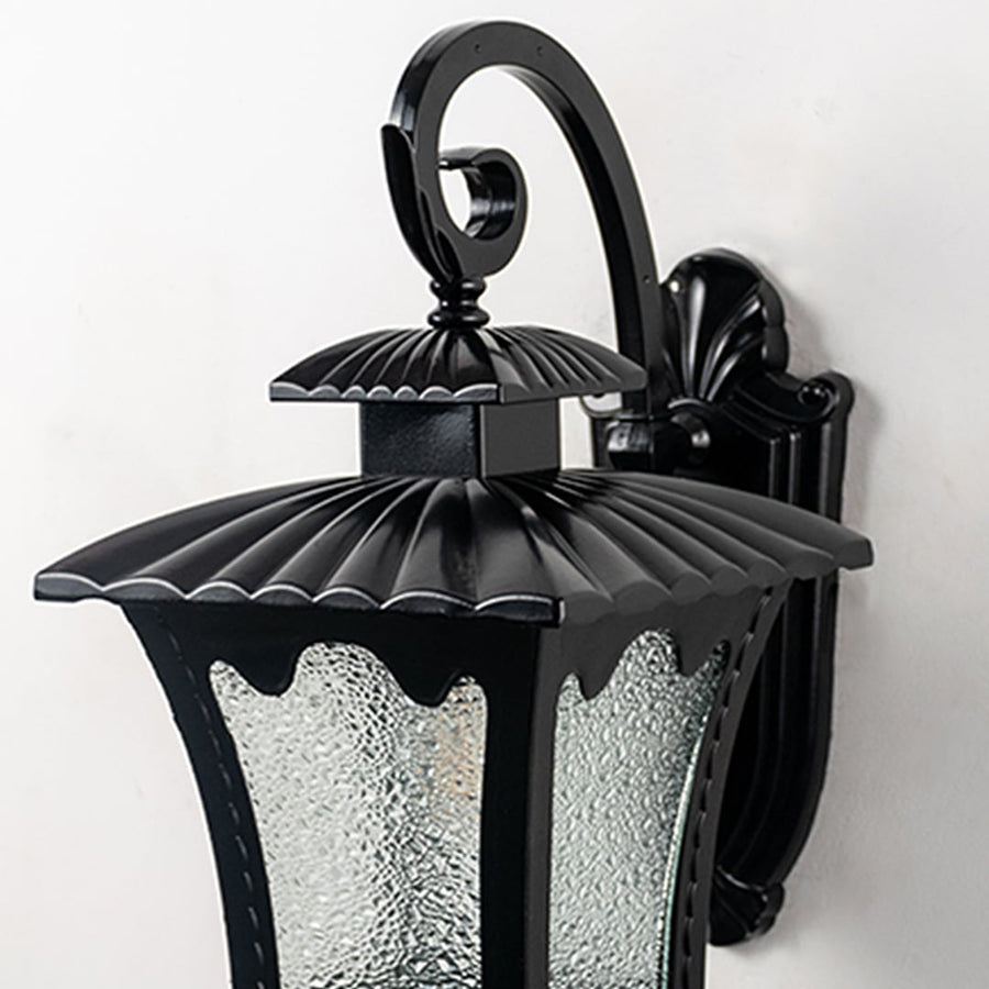 Closeup view of lantern outdoor wall light with curved arm scalloped metal top and frosted glass panels glowing softly.